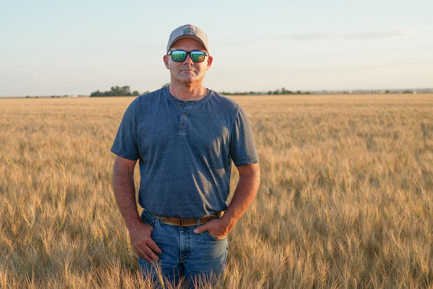 A man wearing a t-shirt, baseball hat and reflective sunglasses stands in a hip-height field of wheat.