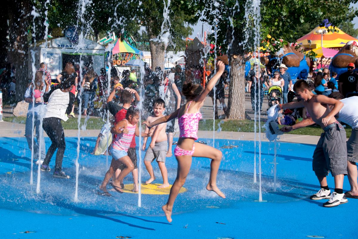 Children dressed in colorful shorts and bathing suites play in fountain sprays at the state fairgrounds in Pueblo, colorado. The area under the fountain is painted blue. There are brightly colored canopies for booths in the background