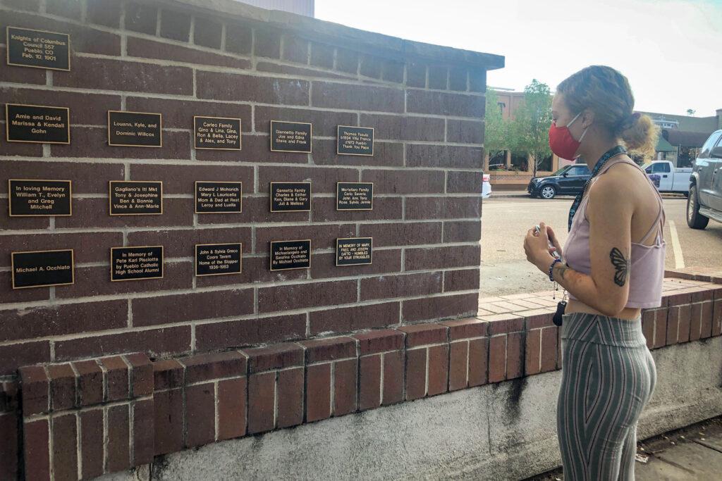 A young woman with a face mask looks at a head high brick wall with small plaques bearing the names of families and individuals.
