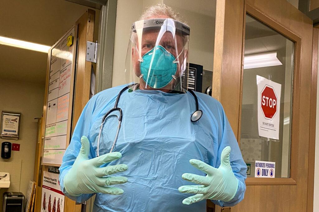 A man stands in a hospital wearing a full range of personal protective equipment including blue gloves, blue plastic gown, a facial barrier mask and a face mask