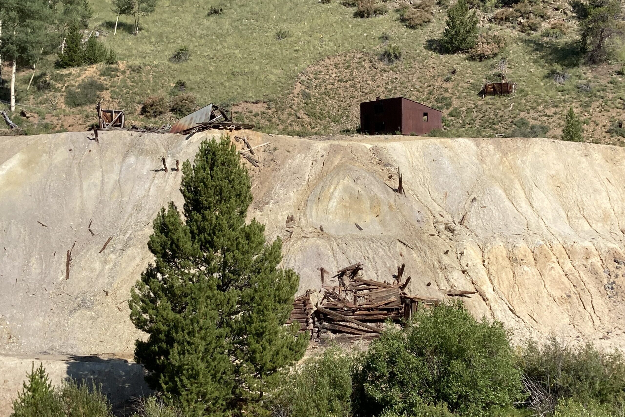 A green hillside is broken by a wall of beige sand with the collapsed remains of old buildings at the top and bottom.