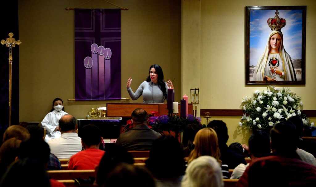 A woman with long black hair stands at the pulpit at a church with an icon of the Virgin Mary behind her on the wall.