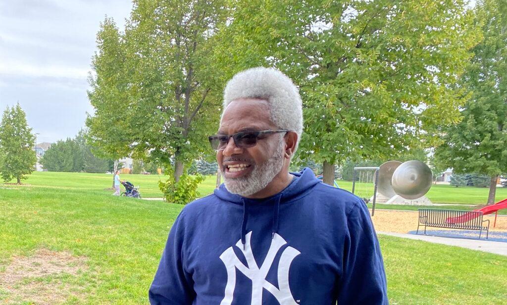 A Black man wearing sunglasses and a blue hoodie stands in a park.