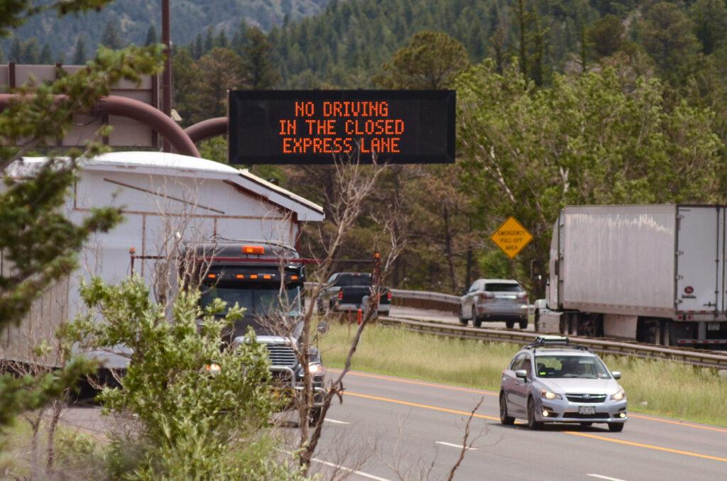 A sign on Interstate 70 west of Idaho Springs warns drivers about toll lane rules. The road has several cars, including two semi's and sedans.