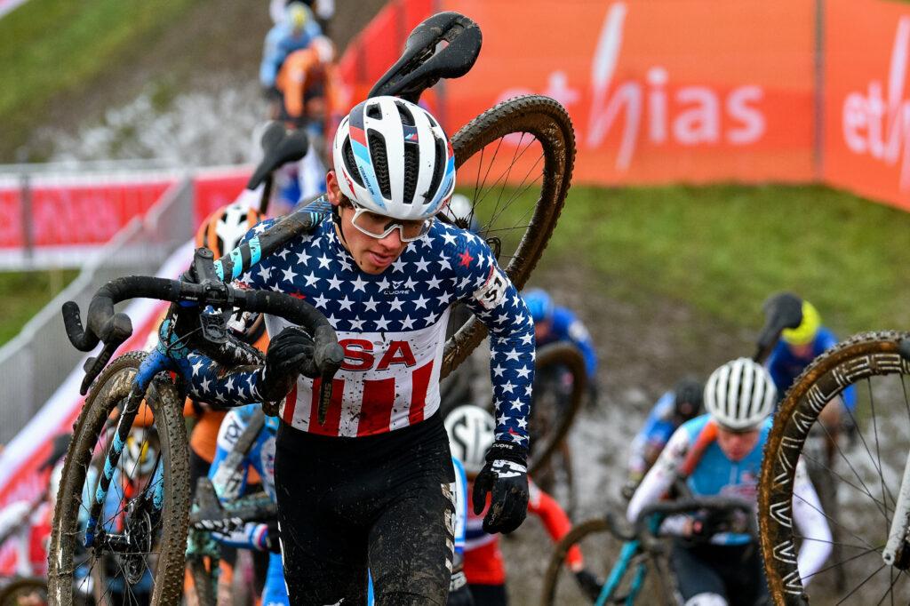 A man carries a bike on his back during a race in a USA bib with competitors around him.