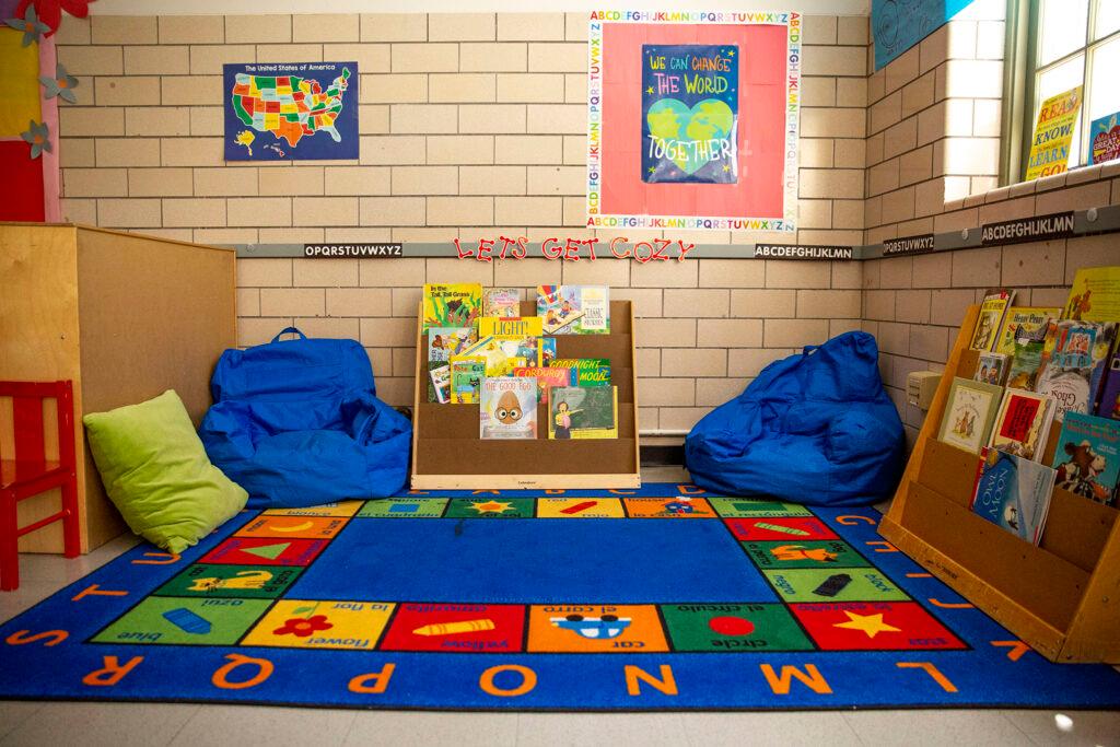 A play area in a child care center. Two blue bean bags leaning up against the brick wall on top of a blue play carpet. There is a bookshelf between the bags. Light from a window to the right seeps in.