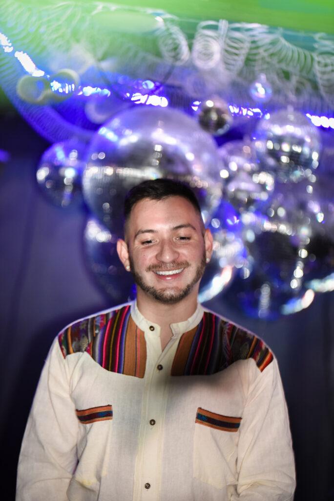 Photo is a headshot of Machu Linea smiling while looking away from the camera. The background features a lot of sparkling disco balls.