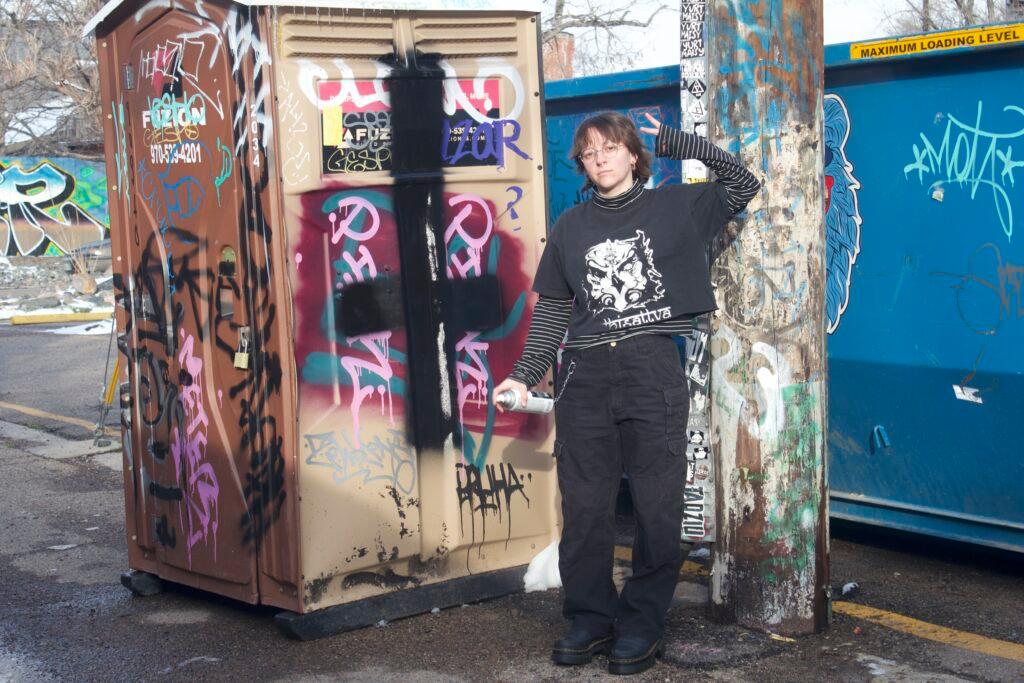 Sam Bosch posing with a peace sign while standing in an alley with a lot of graffiti holding a can spray paint hinting that they too tagged the porta potty.
