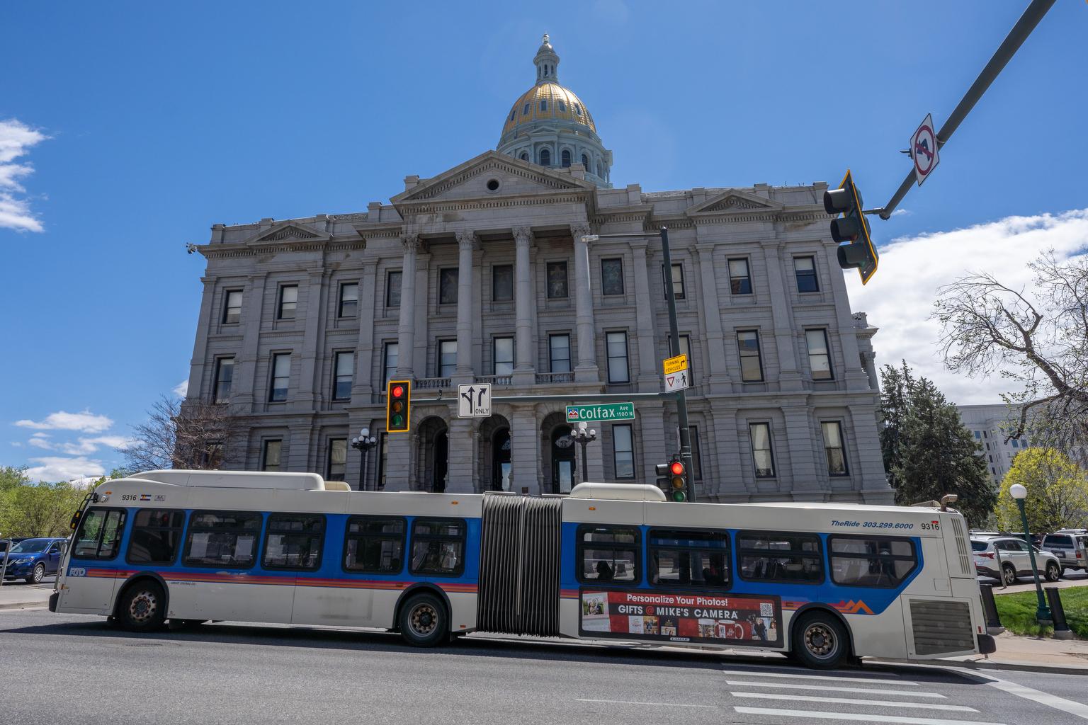 A bus passes in front of the Colorado state capitol building with blue skies in the background.