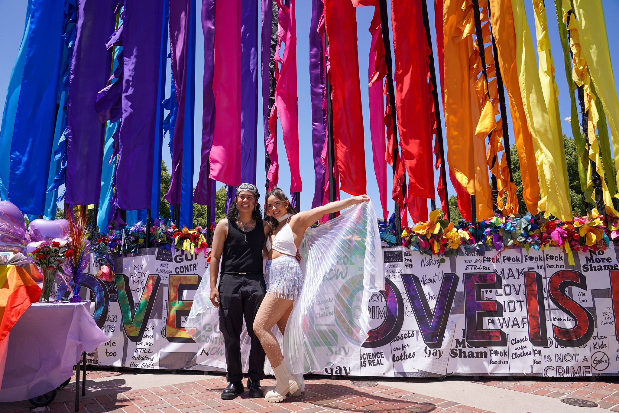 Two people, one wearing a black tank top and black pants with the other wearing white shorts and a white cropped tank top with a white sheer scarf, stands in front of decoration for PrideFest.