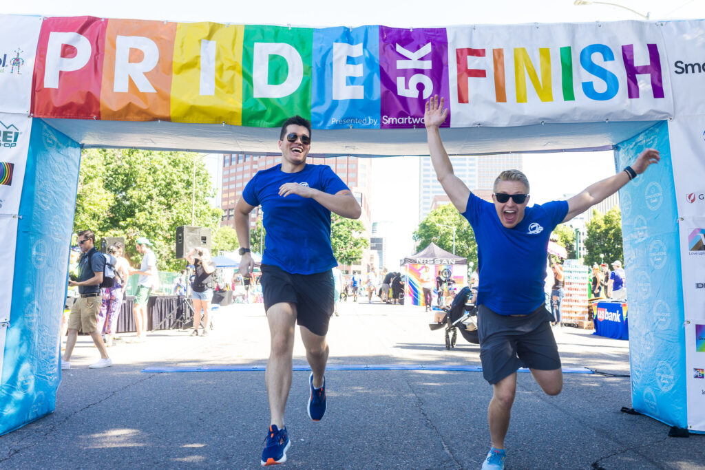 Two people wearing matching blue shirts and grey cargo shorts and sunglasses run under an inflatable archway with a banner reading Pride 5k Finish.