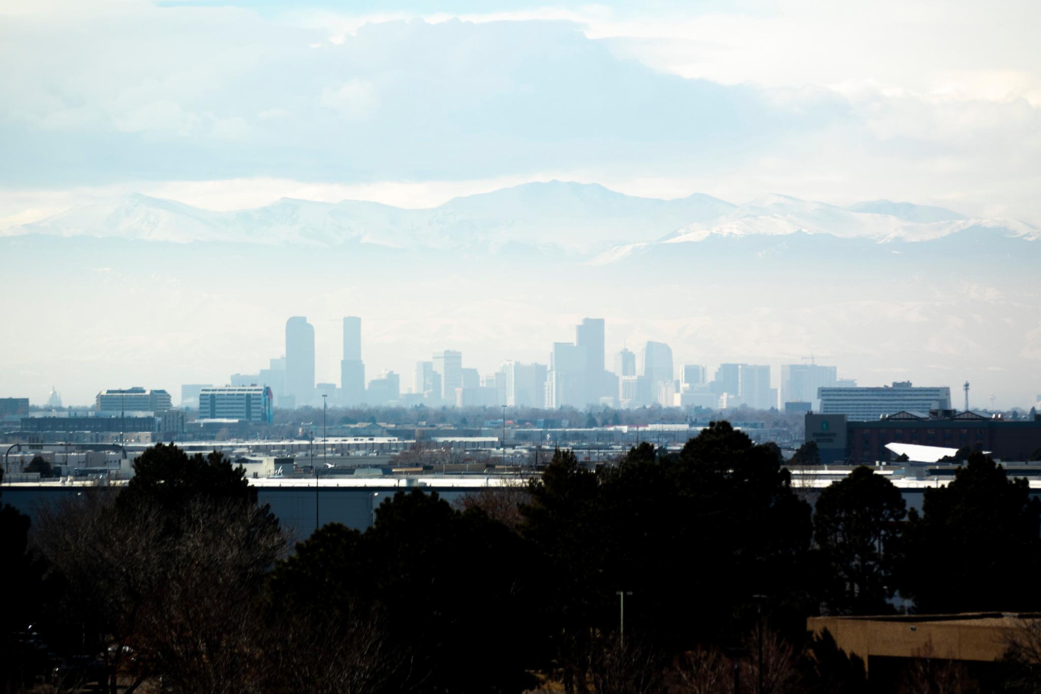 The downtown Denver skyline seen from far away — the buildings nearly match the color of the mountains and clouds behind them, melting together from all the hazy air between them and the camera.