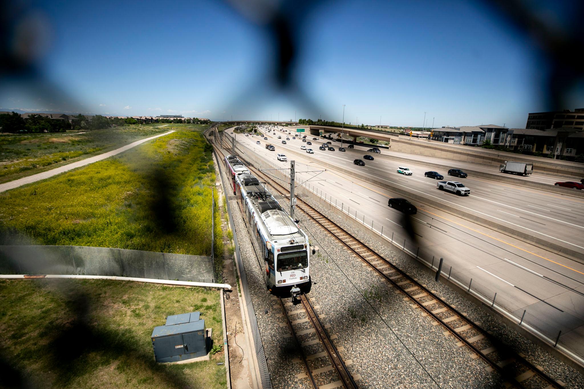 View of an RTD train through a chain link fence that runs parallel to I-25.