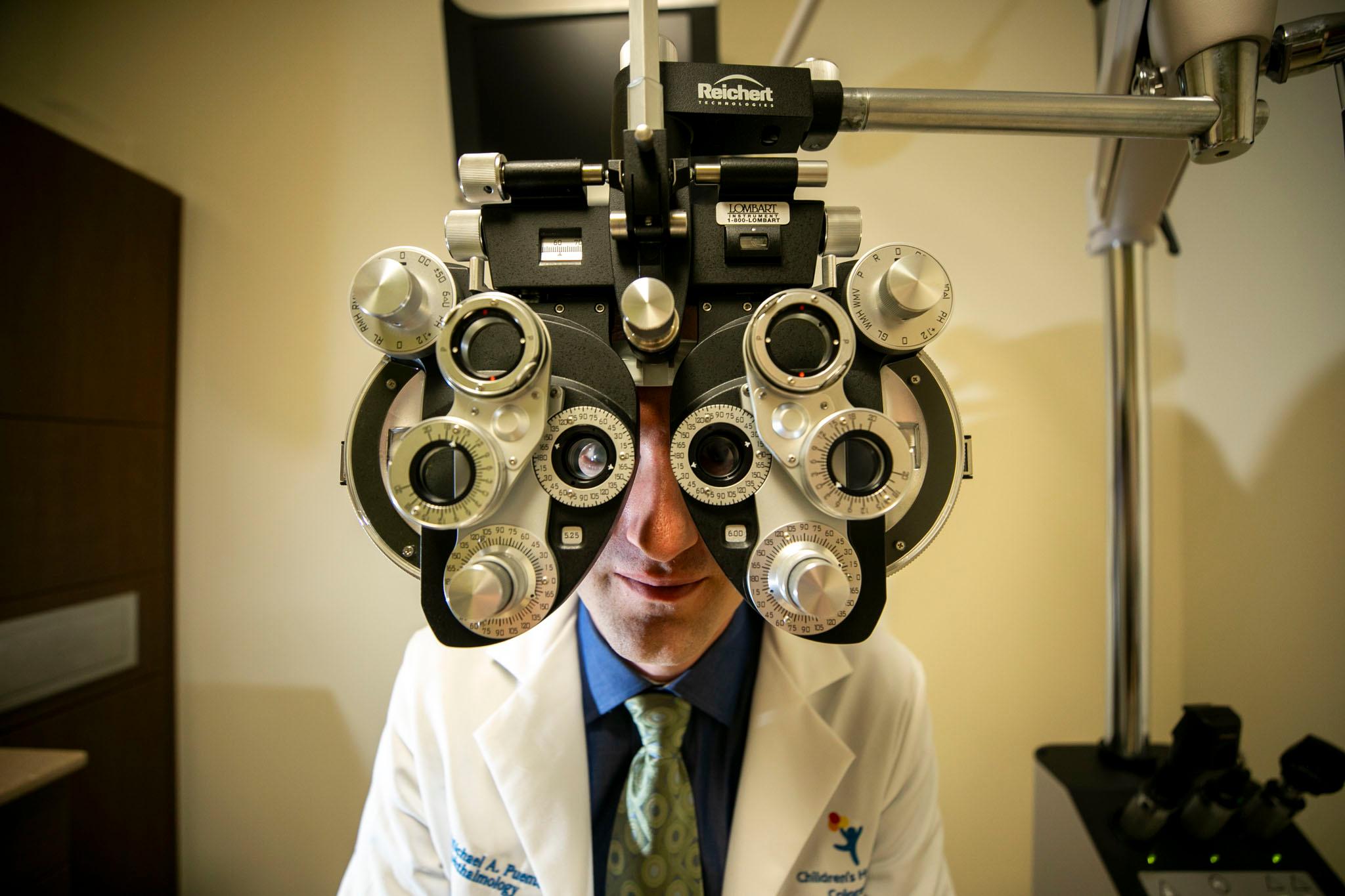 A man wearing a white lab coat and a blue button down shirt peers into a medical device used by eye doctors at the camera.