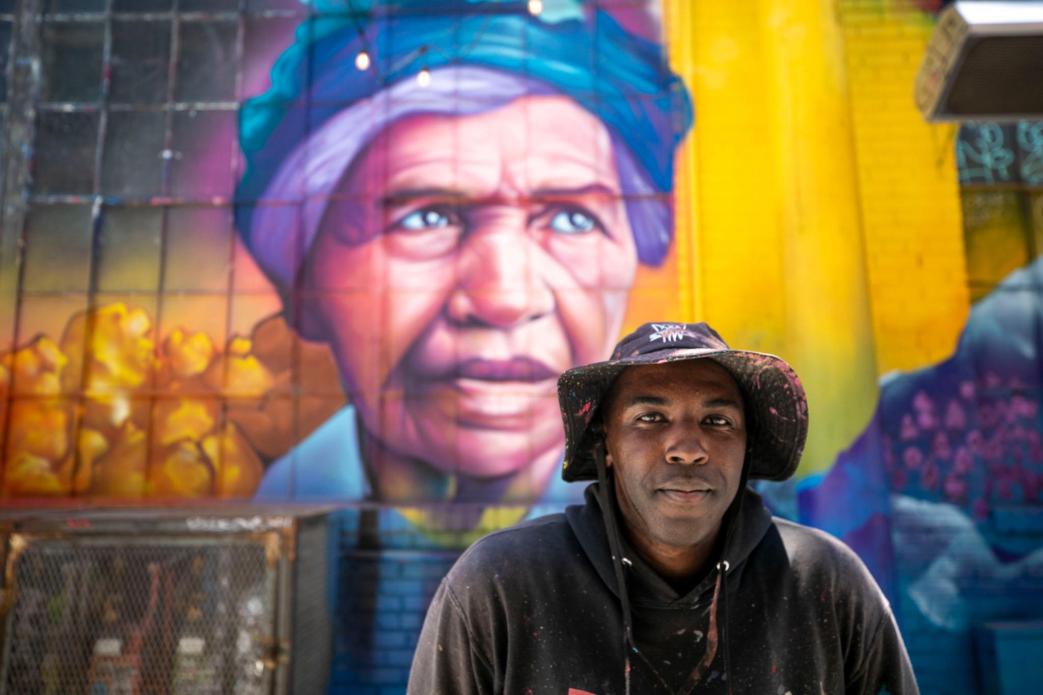 Artist Detour stands in front of a new mural he painted depicting depicts Clara Brown in vibrant colors