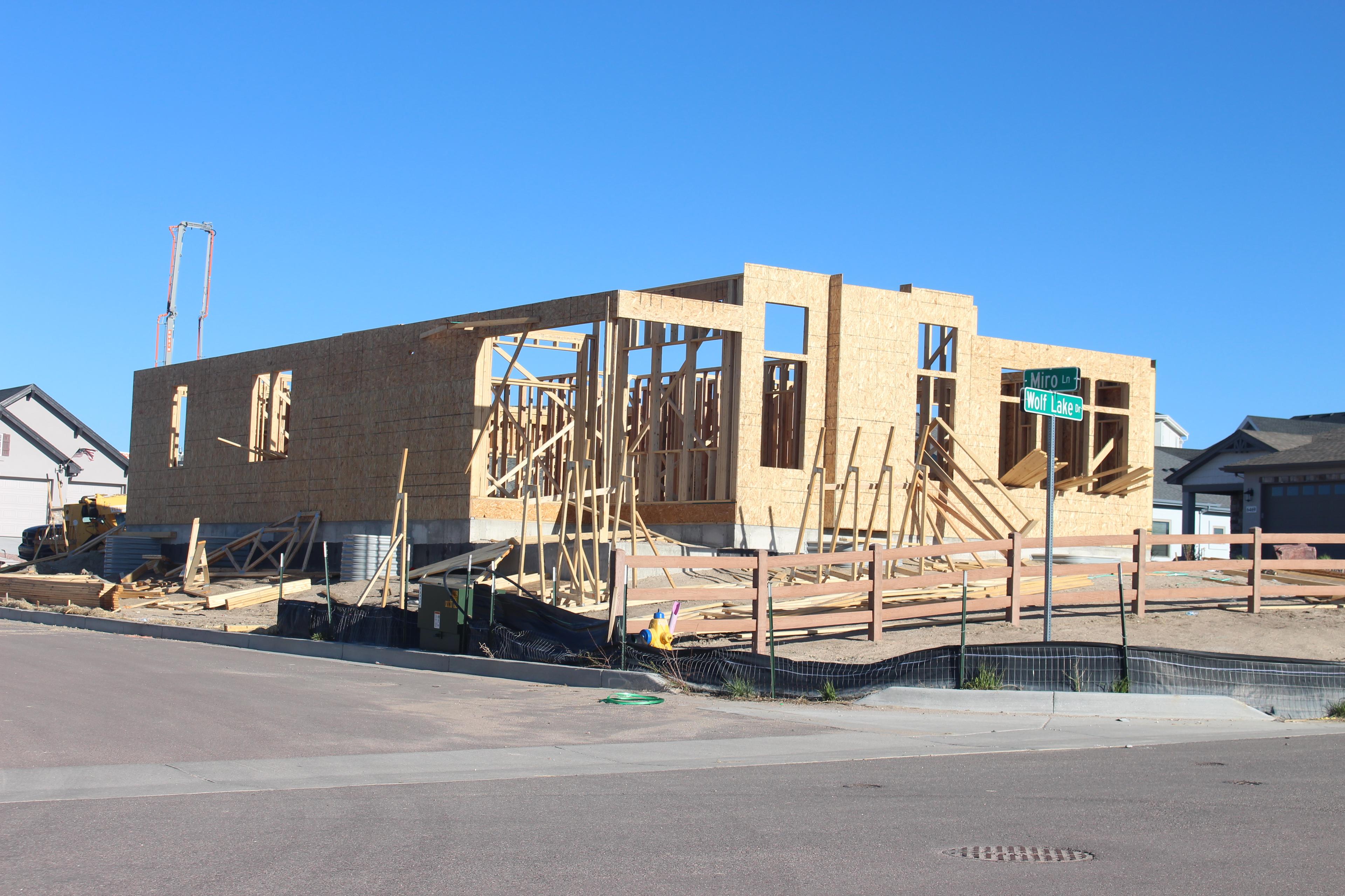 a partially constructed home with parts of the OSB walls standing but no roof yet. The framing wood is up for the interior walls. a street sign shows the house is at the corner of two paved streets called Miro lane and Wolf Lake Dr. The sky is blue.