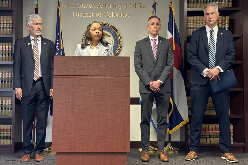 A woman speaks from a podium at the U.S. Attorney's Office for the District of Colorado. Three men in suits stand behind her.