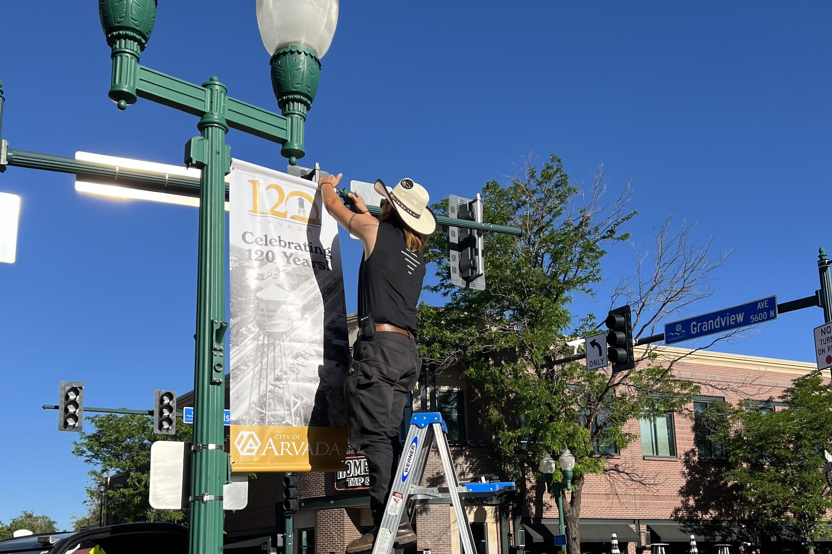 A worker hangs a banner proclaiming Arvada's 120th anniversary from a lightpost, June 2024.