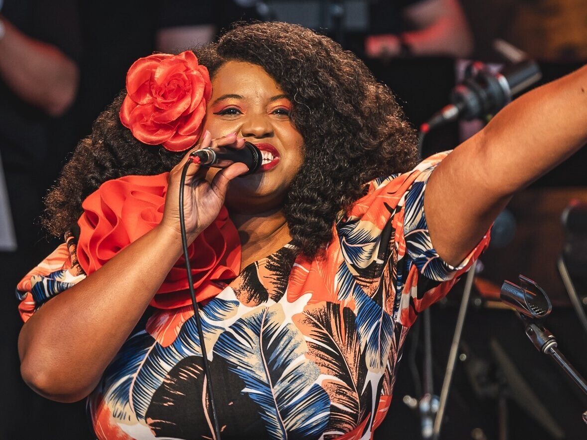 A woman wearing a colorful dress and a flower in her hair holds a microphone and sings into it.