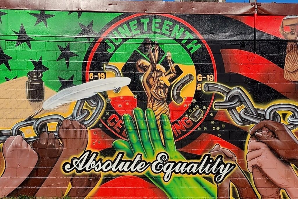 Mural on a brick wall predominantly using the colorso black, red, yellow, and green. Hands are reaching up from below to break chains, there are historical figures on the right and left.