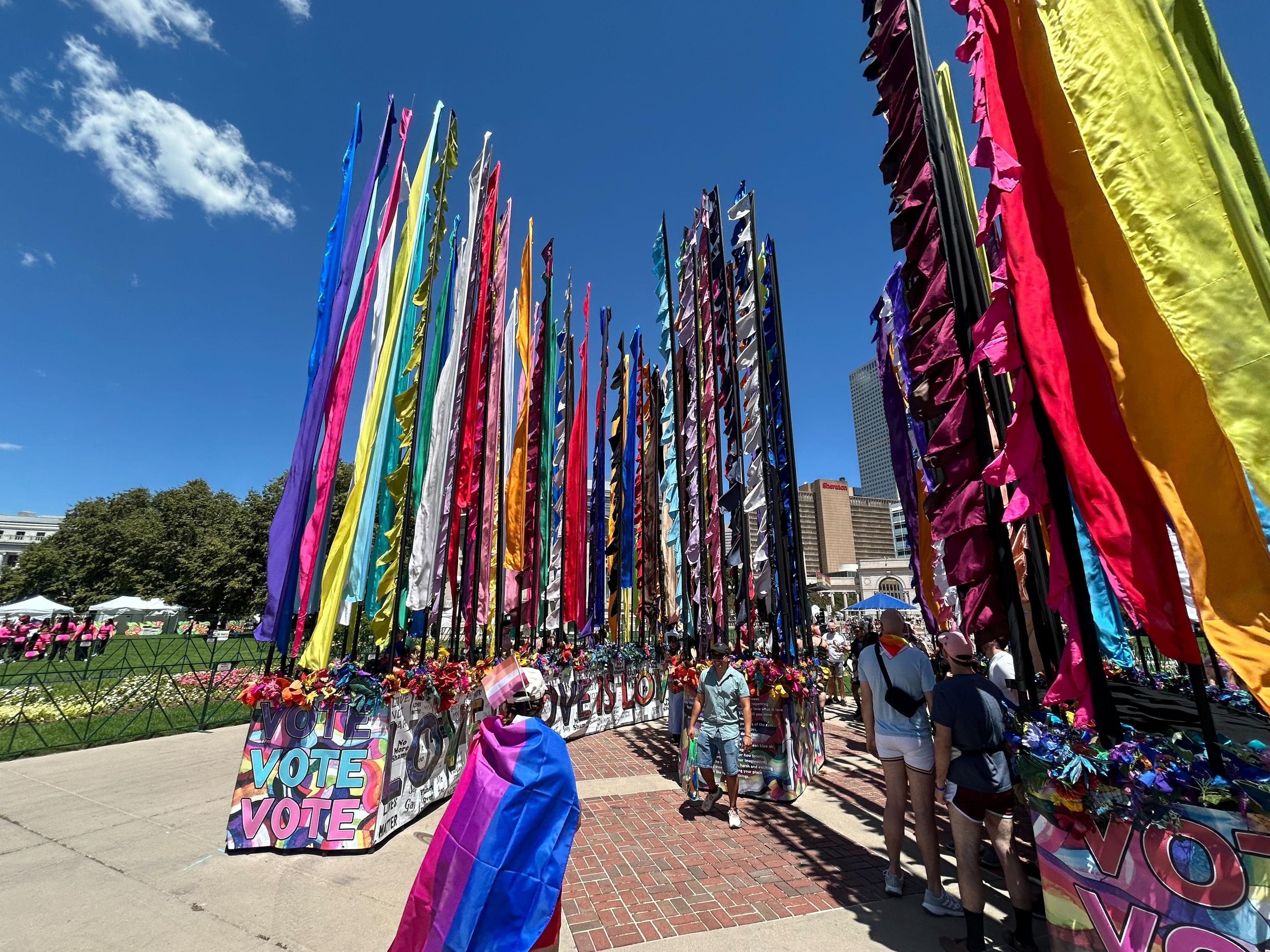 People walk among a sculpture featuring multicolored flags that rise high in the air and flowers.