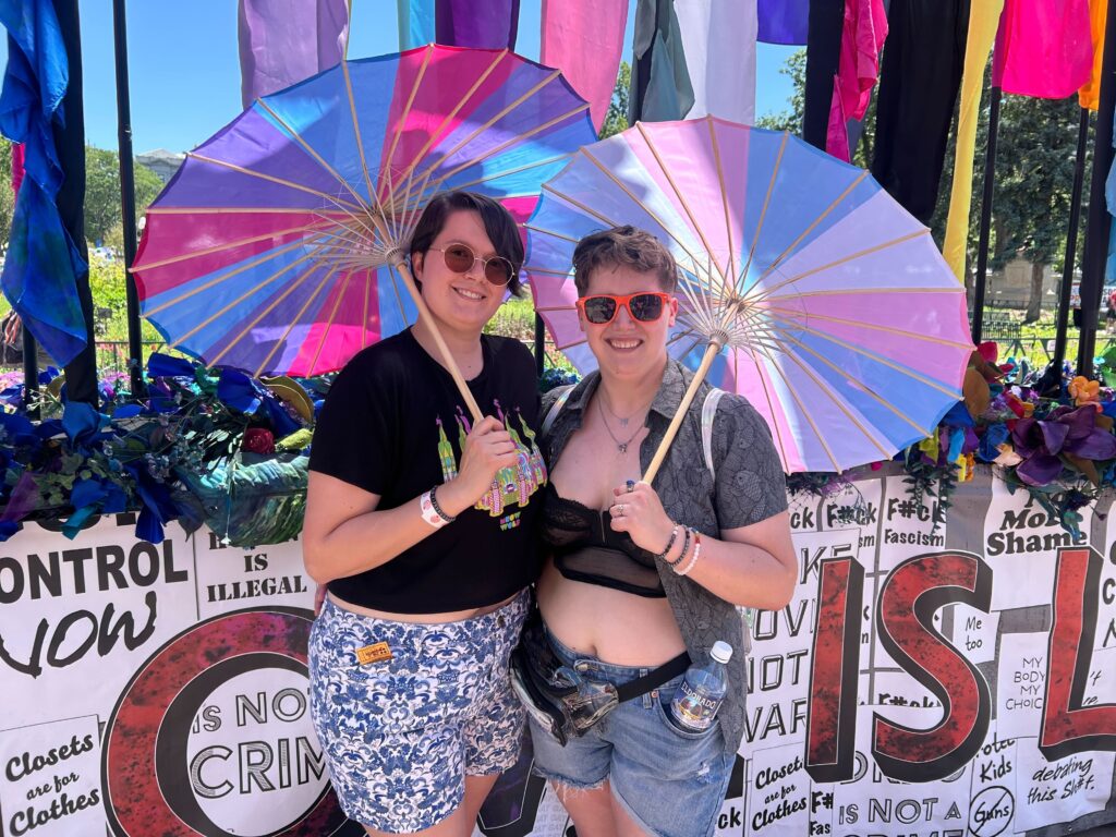 Two people hold pink, purple and blue umbrellas as they stand underneath a sculpture featuring multicolored flags that rise high in the air and flowers.