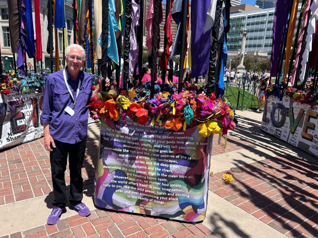 A man poses with a sculpture he built featuring multicolored flags that rise high in the air and flowers.