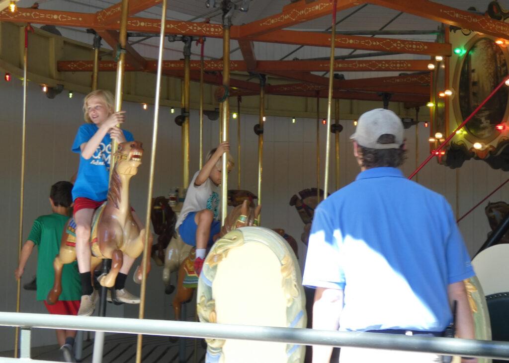 A child sits on a horse on a carousel while a man in a blue short sleeve button-down shirt and a ballcap looks at him.