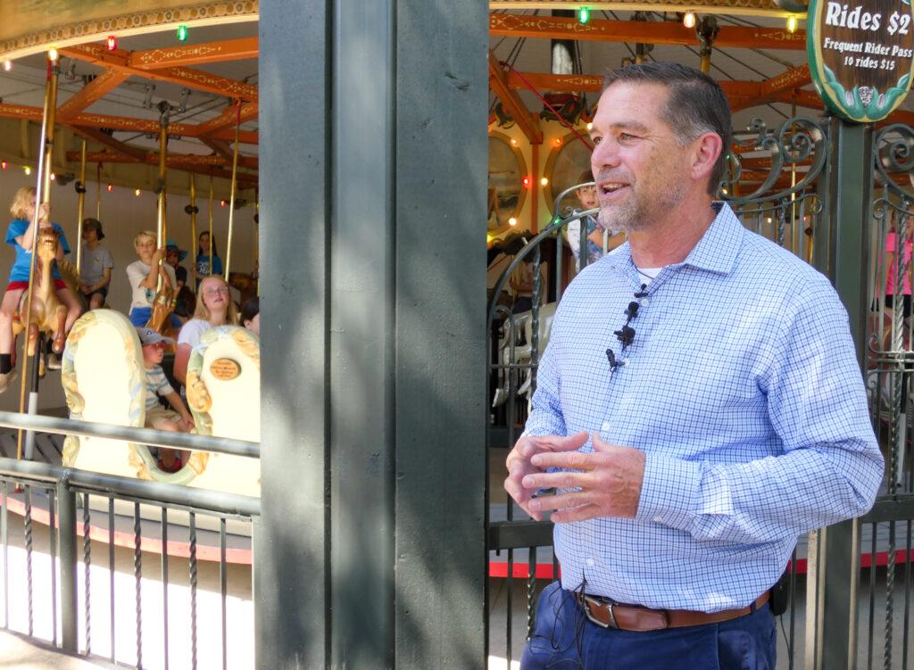 Man in a blue button down shirt stands with his hands in front of his stomach pressed fingertip together.