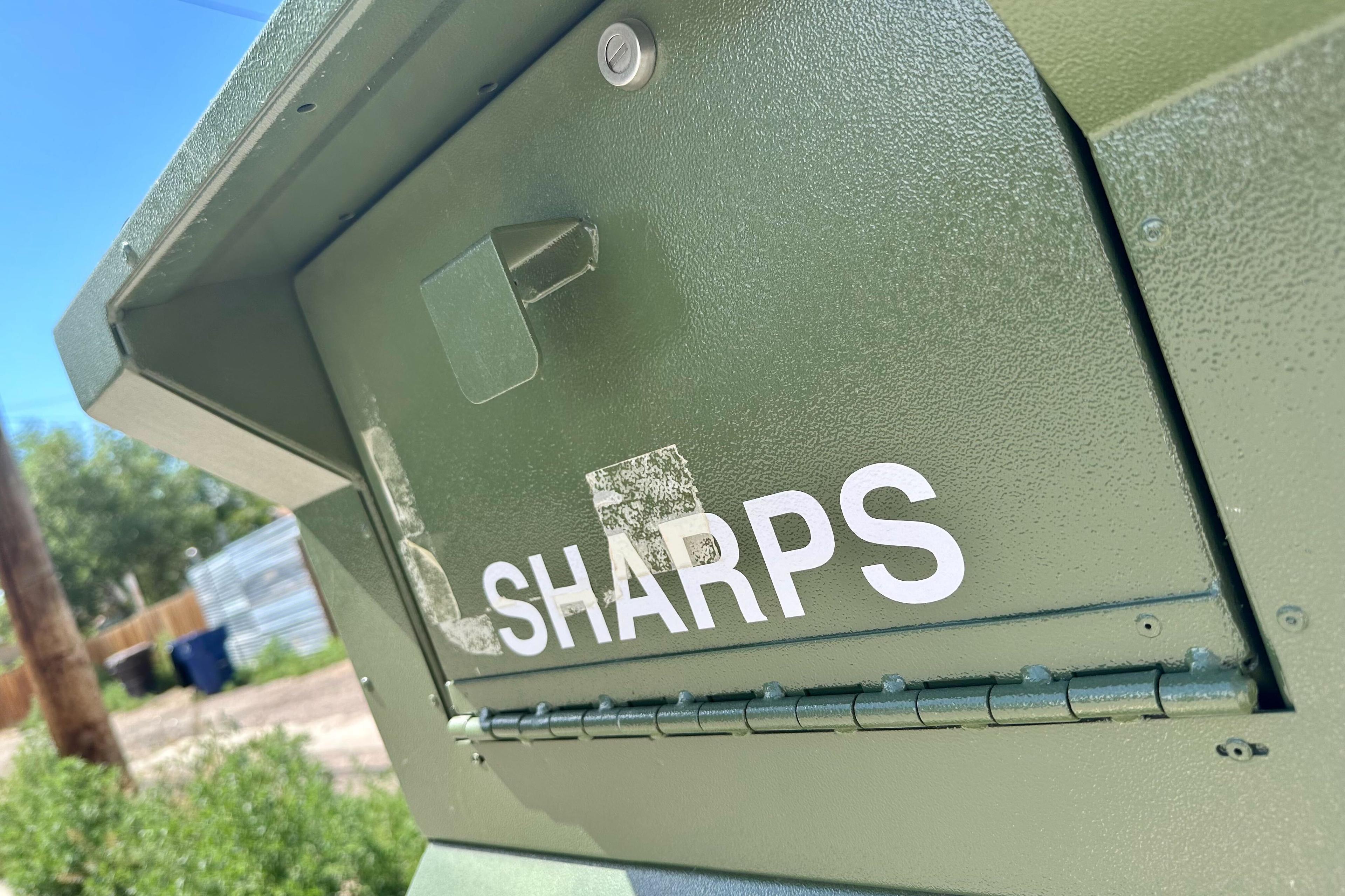The door of a green metal sharps container box is shown under the midday sun.