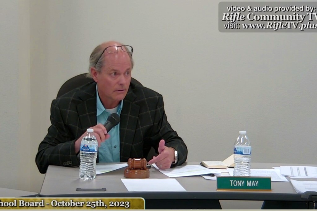 Man in a blue button down shirt and black stripped jacket holding a microphone at a table with two water bottles and papers strewn about. His name tag, Tony May, is sitting on the table.