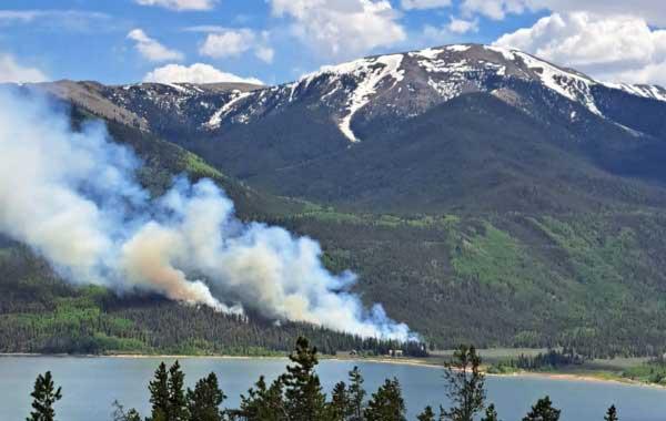 Visible smoke from a fire burning through the pine forest surrounding an alpine lake.