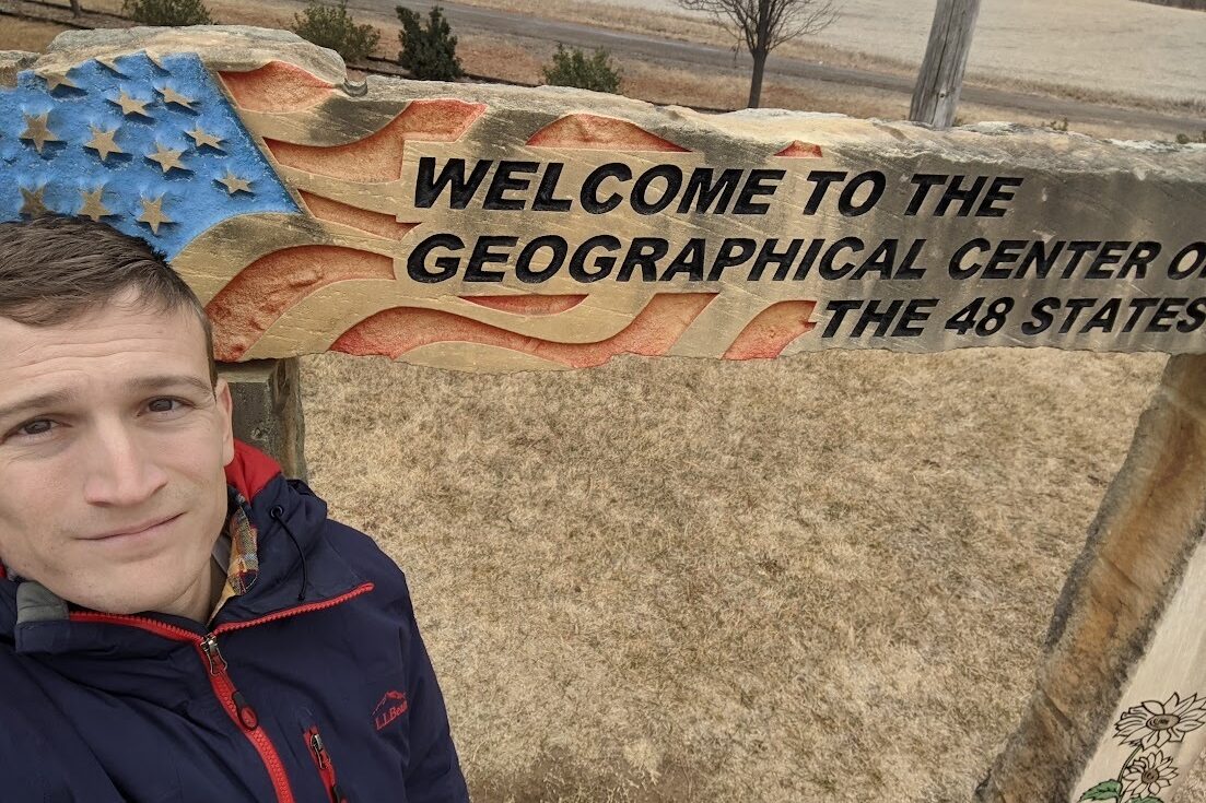 Man in a navy zip up jacket with red trimming takes a selfie looks up at the camera in front of a sign.