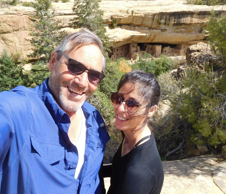 A man wearing a blue button down shirt, white undershirt, and sugnalsses smiles at the camera while a woman with sunglasses and a black shirt stands facing him with head turned toward the camera. There is a desert scenery behind them.