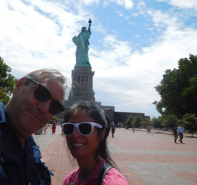 Two people stand in front of the Statute of Liberty. A man in a blue button down shirt and sunglasses leans down near a woman wearing sunglasses, a pink shirt, and a backpack.