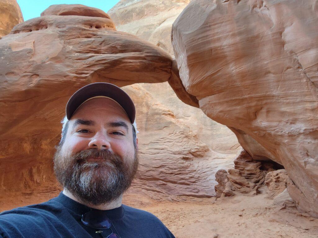 A man with a beard wearing a hat smiles at the camera with rock formations behind him.