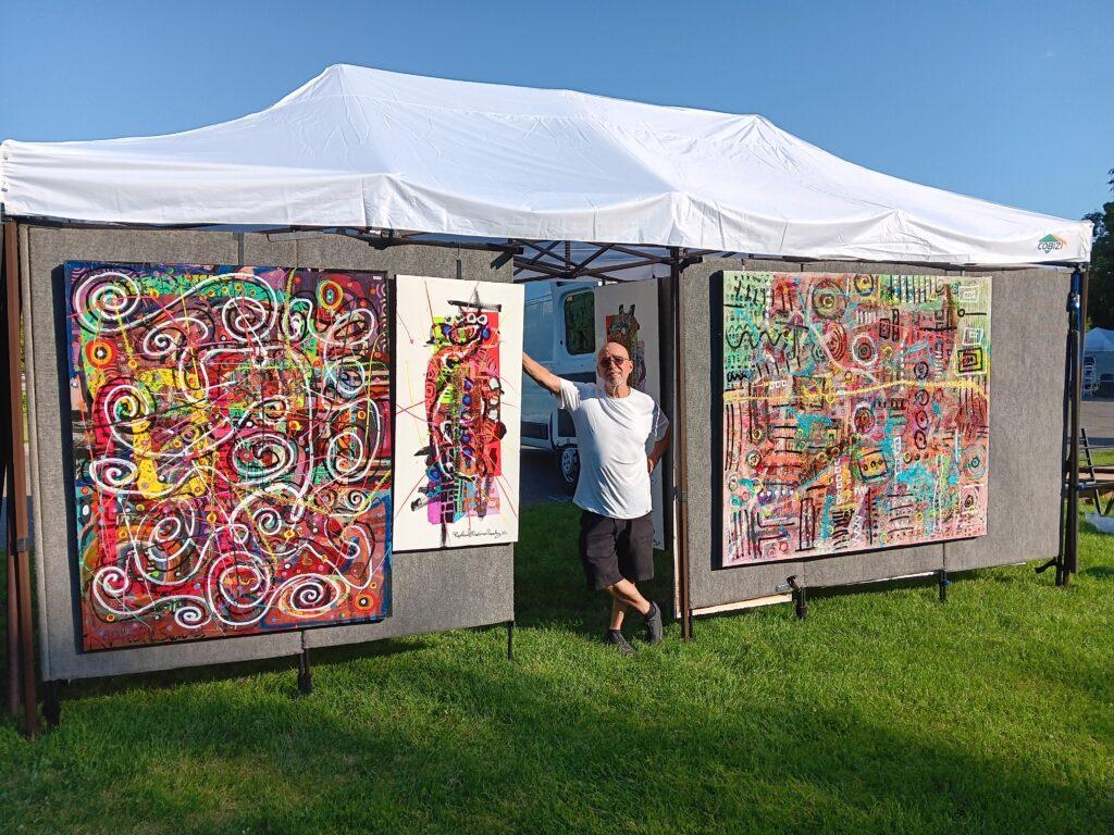 A man in a white t-shirt stands in a tent surrounded by multicolored artworks.