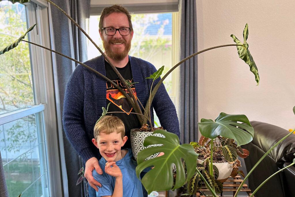 A man with a red beard stands behind a small boy. The man is holding a large plant.