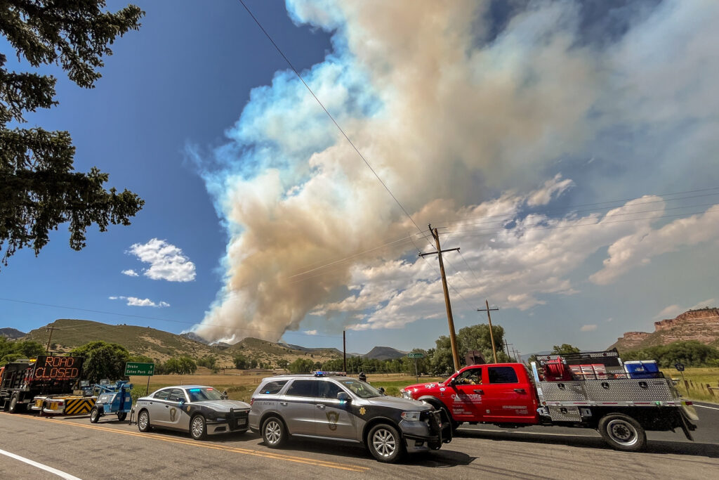 Emergency vehicles sit near a fire burning with wildfire smoke visible in the background.