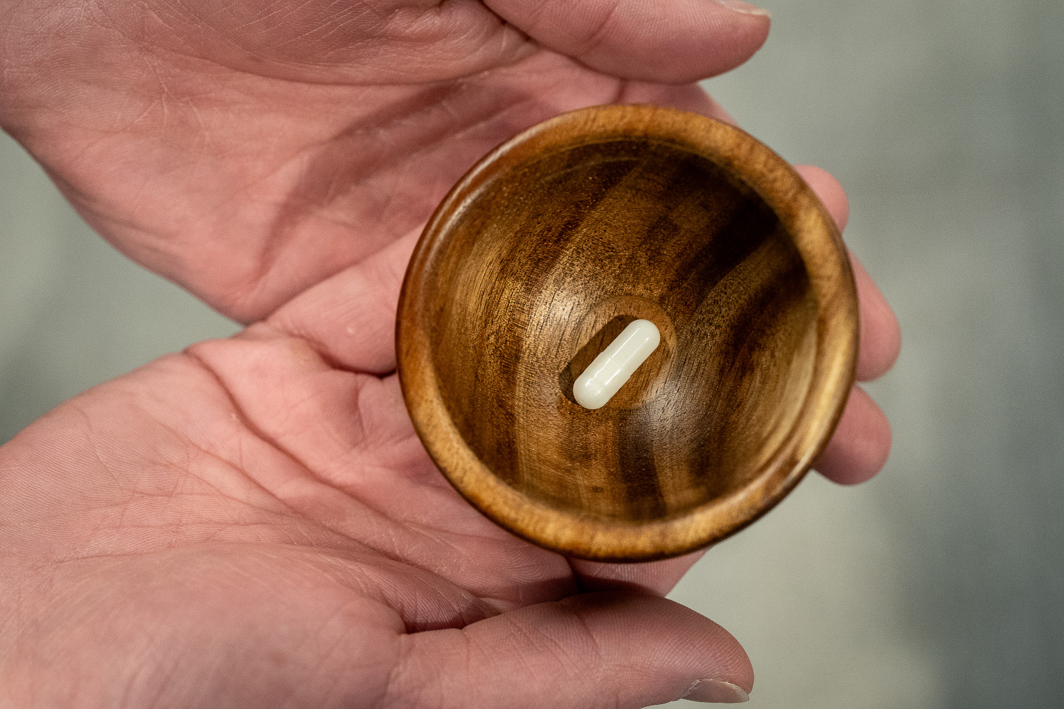 Two hands cup together to hold a wooden tiny round bowl that has a single white pill in it.