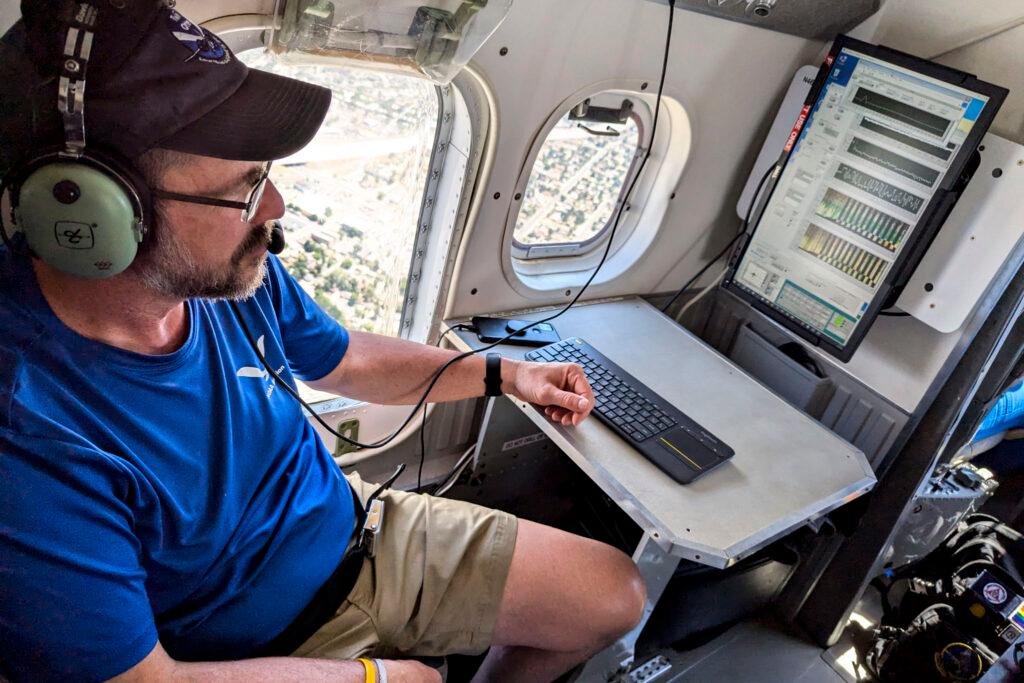 Alan Brewer wears a blue shirt khaki shorts with a black hat glasses and headphones in the cockpit of an airplane.