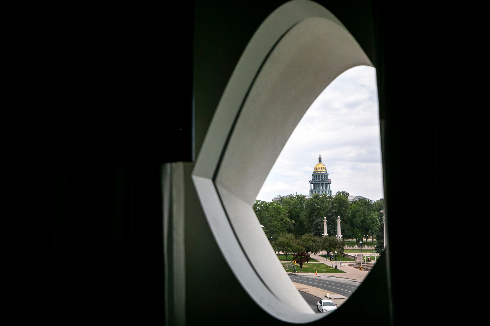 The Capitol's gold dome stretches over green trees, seen through an eyeball-shaped window; the room around the window is black.