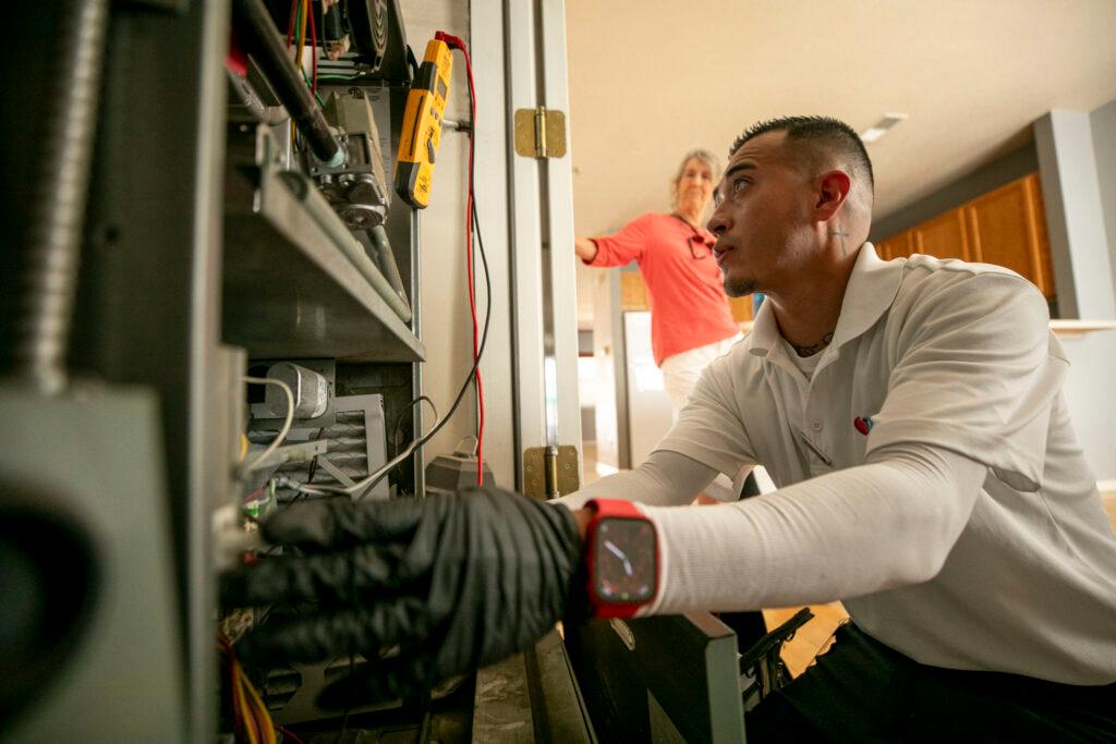 Man works an air conditioner unit inside a home while a woman watches at a distance.