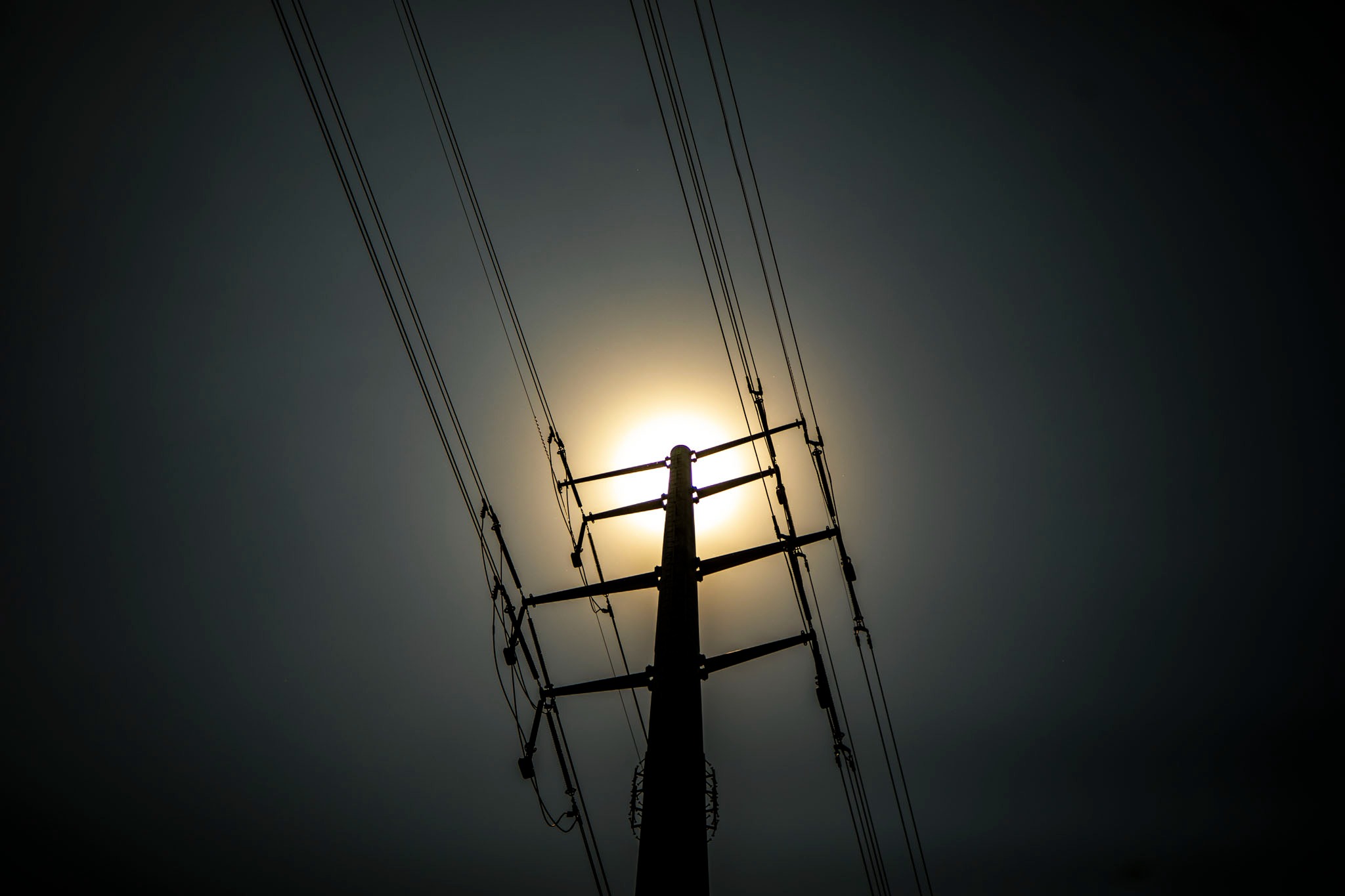 The shape of a utility pole is silhouetted by a bright orange sun.
