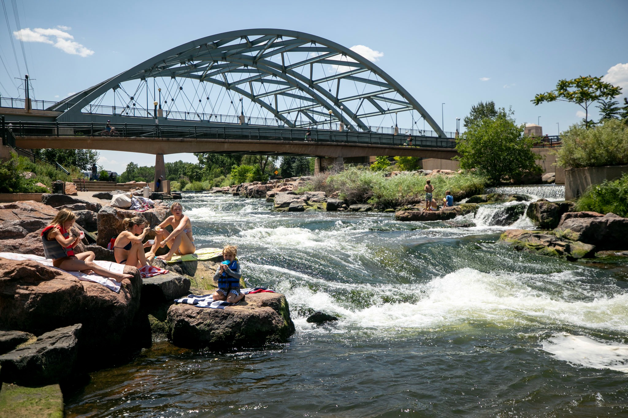 People in swimsuits sit on rocks next to a flowing rapid. A big metal bridge looms behind them.