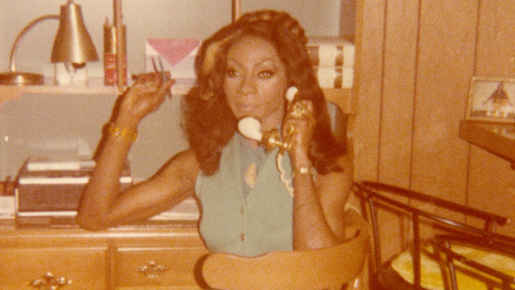 A woman sits sideways on a wooden chair holding an old wired phone in one hand against her ear the other a pen. She's wearing a blue sleeveless garment with buttons and a high neck underneath it. There is wood paneling and a shelving unit behind her.