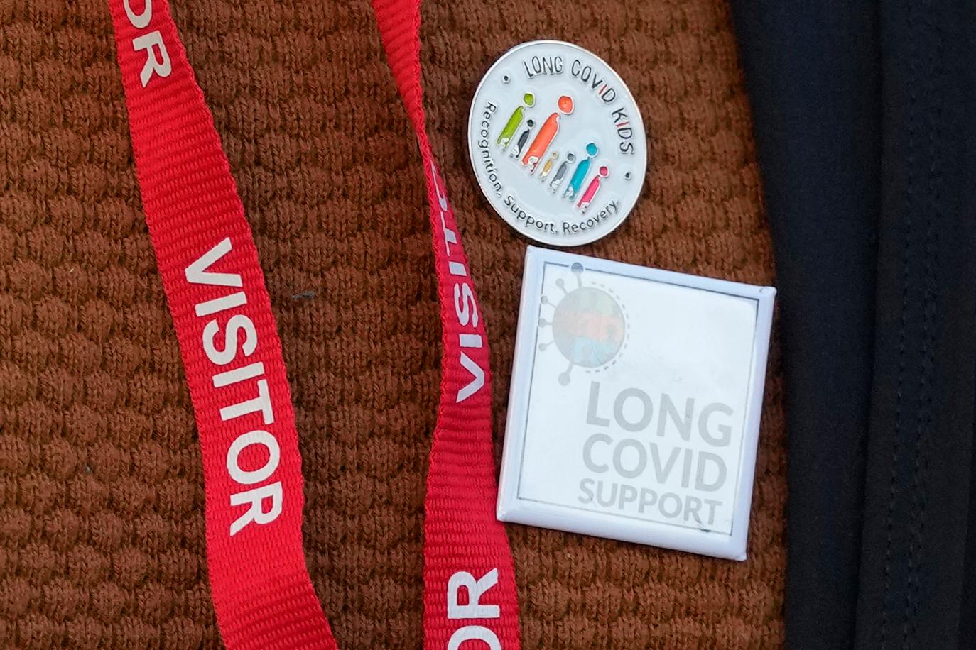 A close-up image of an array of buttons and a lanyard on a person's orange knit shirt. The lanyard is red with "VISITOR" on it. Both buttons are in support of long COVID treatment.