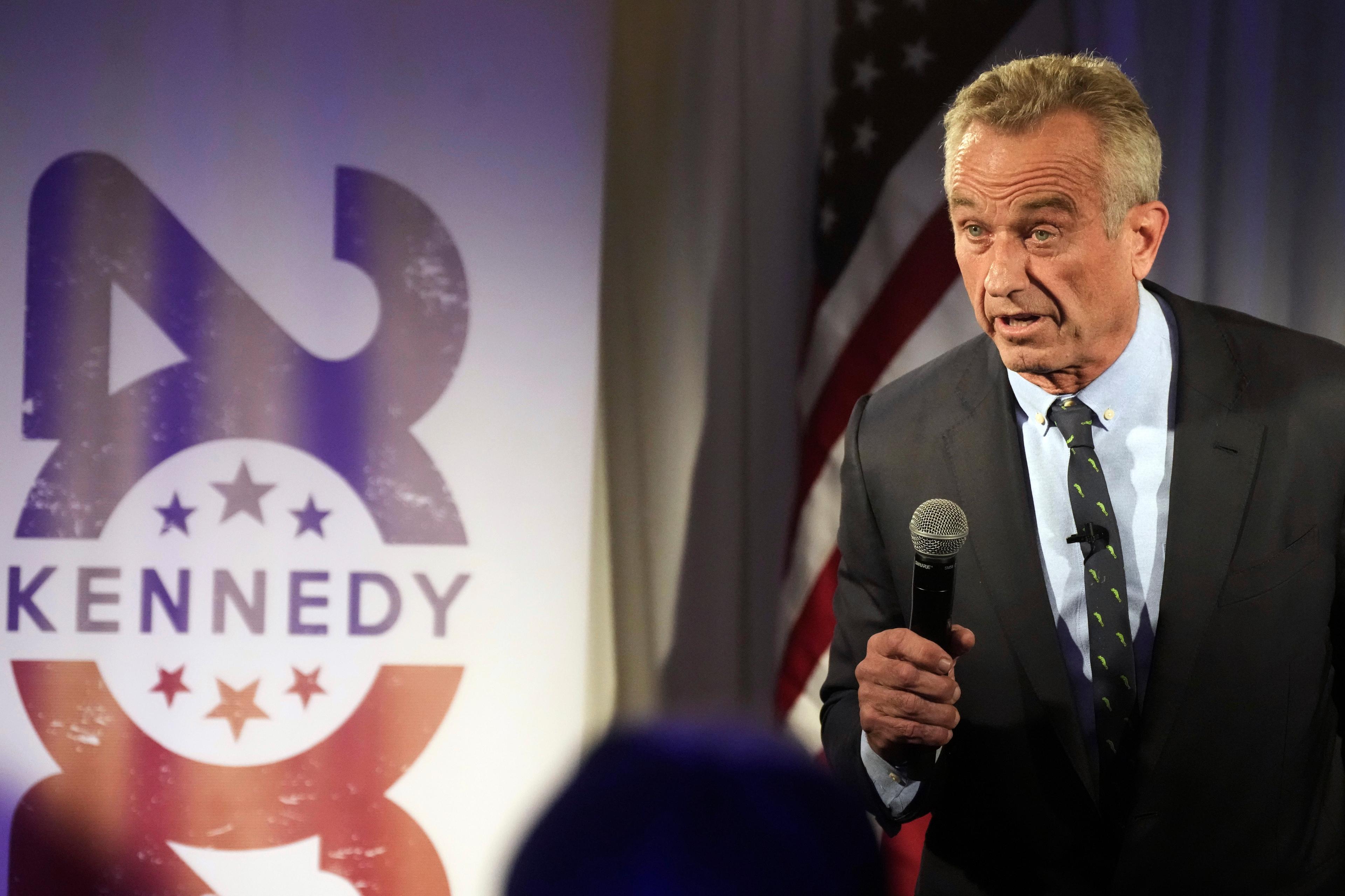 Robert Kennedy Jr. stands with a microphone in his hand near a sign in the background that promotes his campaign.