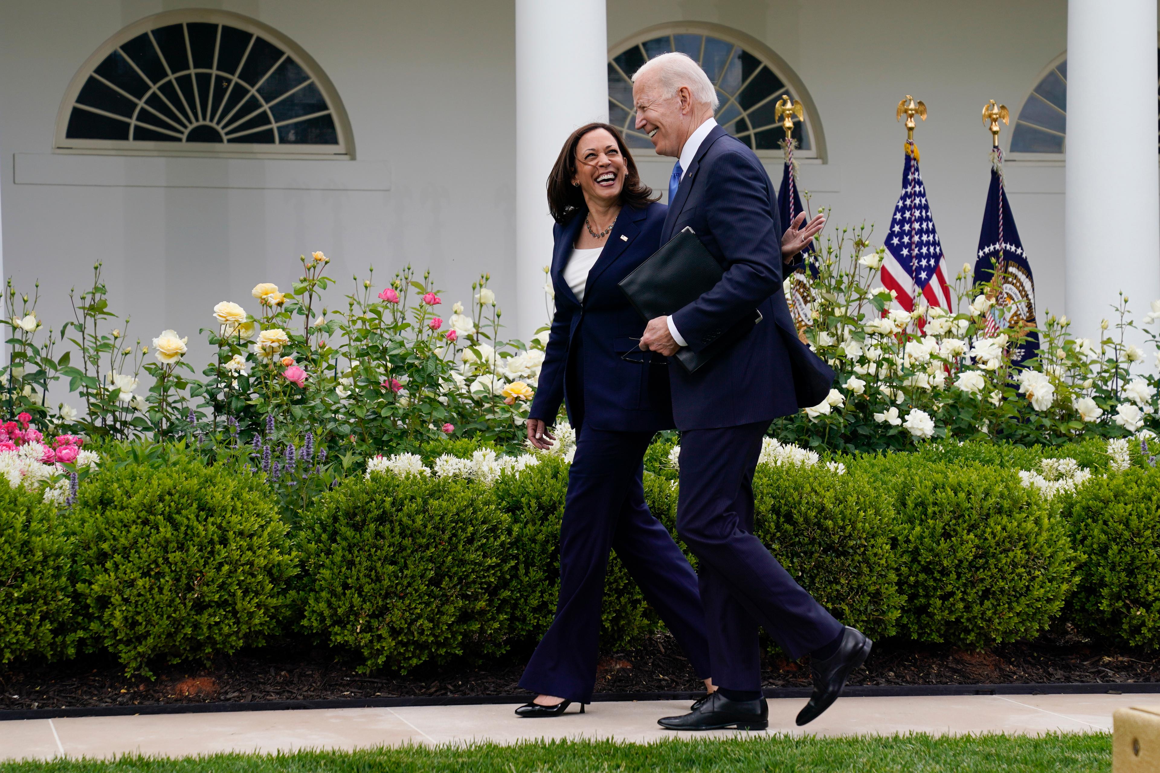 Kamala Harris pats Joe Biden on the back as the two walk at the White House Rose garden with bushes and flowers in the background.