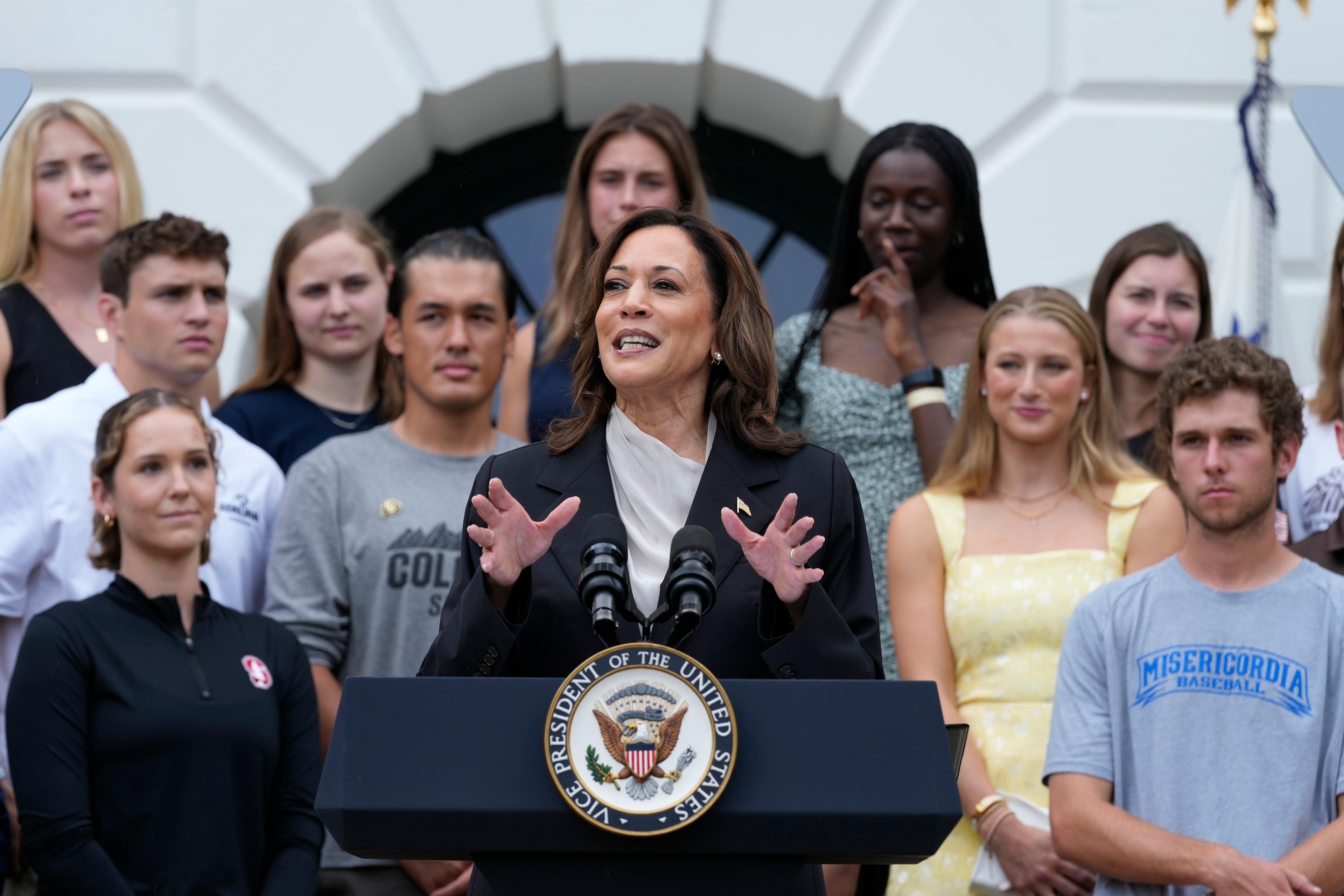 Vice President Kamala Harris speaks at a podium on the South Lawn of the White House at a navy podium with the President of the United States seal on front with two mics attached. Behind her are 11 visible college athlete. The White House is barely visible behind the students.